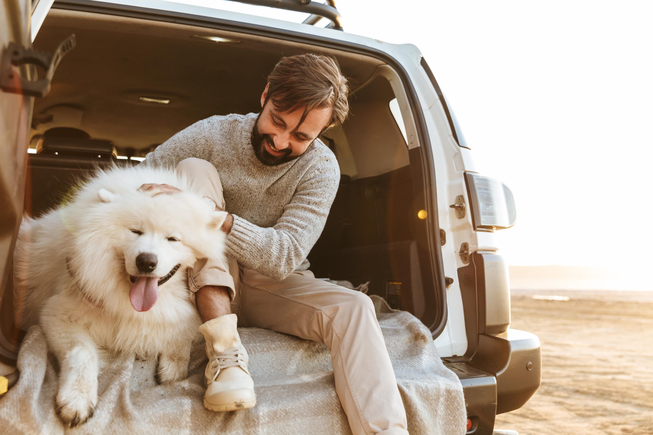 Ein junger Mann sitzt mit seinem weißen Hund im Kofferraum eines Autos. Er sieht glücklich aus und streichelt seinen Hund.
